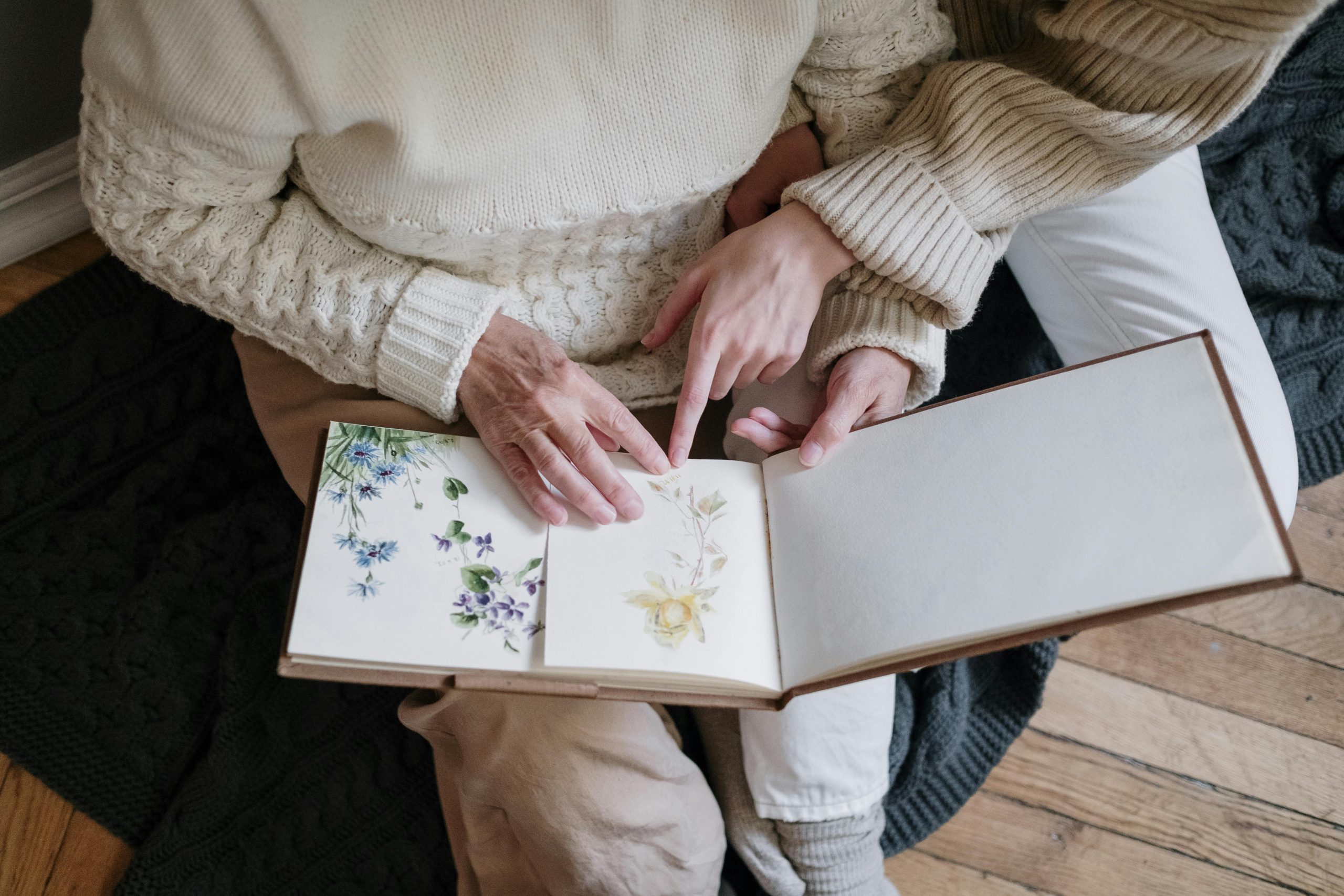 Two women reviewing a family photo album with floral artwork, creating memories together.